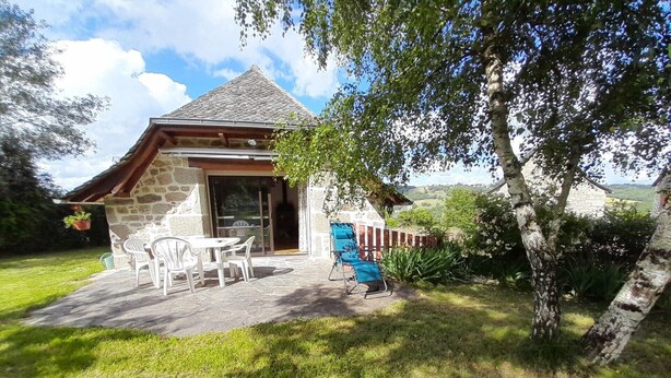 Une maison en pierre avec un toit de tuiles, une terrasse avec des chaises et une table en plastique blanc, et une vue sur la campagne.