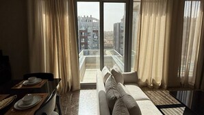 Een moderne woonkamer met een bank, eettafel en een stadsgezicht door grote ramen.