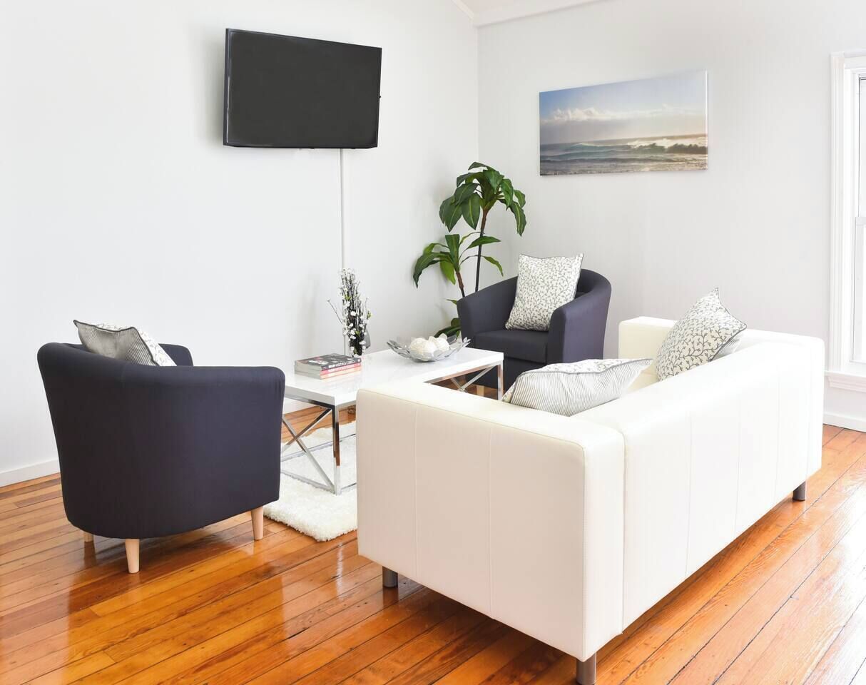 A modern living room with a white sofa, a black armchair, and a coffee table. A flat-screen TV is mounted on the wall, and there is a window with a view of the ocean.