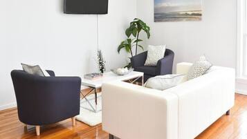 A modern living room with a white sofa, a black armchair, and a coffee table. A flat-screen TV is mounted on the wall, and there is a window with a view of the ocean.