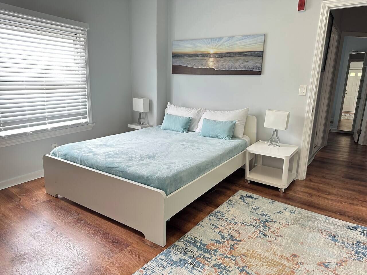 Un dormitorio con cama, mesitas de noche, una ventana con persianas, un cuadro en la pared y una alfombra.