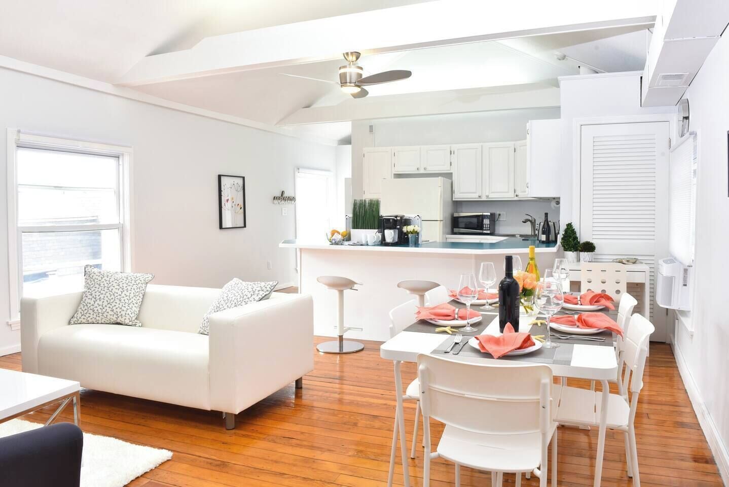 Una sala de estar moderna con un sofá blanco, una mesa de comedor preparada para cuatro comensales y una cocina al fondo.