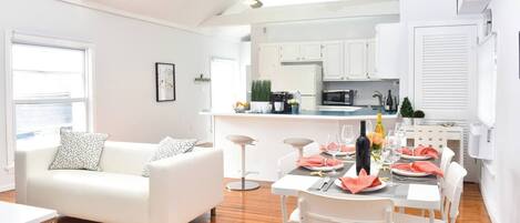 Una sala de estar moderna con un sofá blanco, una mesa de comedor preparada para cuatro comensales y una cocina al fondo.