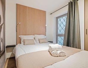Ein Hotelzimmer mit Bett, Kopfstütze, einem Fenster mit Vorhängen und einem Handtuch auf dem Bett.