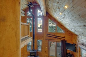 A wooden cabin interior with a loft bedroom, a fireplace, a television, and a deck with outdoor seating. - Beach & Dock Access! Waterfront Payette Lake Home (McCall)