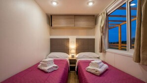 Une chambre double avec deux lits, une table de chevet avec une lampe, une fenêtre avec des rideaux et une vue sur l’extérieur.