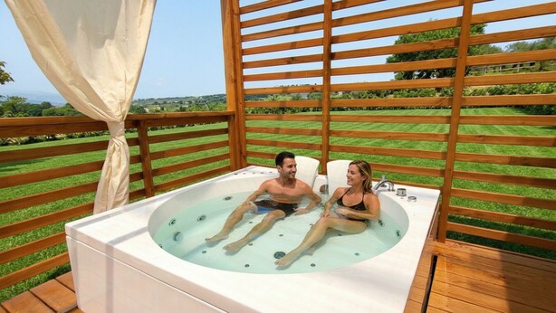 Un couple se détend dans un jacuzzi sur une terrasse, avec vue sur un paysage verdoyant.