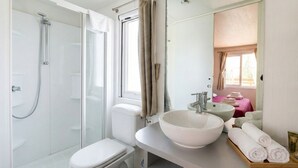 Une salle de bain moderne avec une douche à parois de verre, des toilettes blanches, un grand lavabo blanc et un miroir qui reflète une serviette rose et une fenêtre.