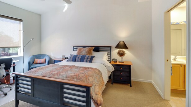 A bedroom with a bed, a desk, a chair, a ceiling fan, a nightstand with a lamp, and a bathroom visible through an open door. - Golf, Hike & Unwind: Oro Valley Townhome w/ Patio! (Oro Valley)