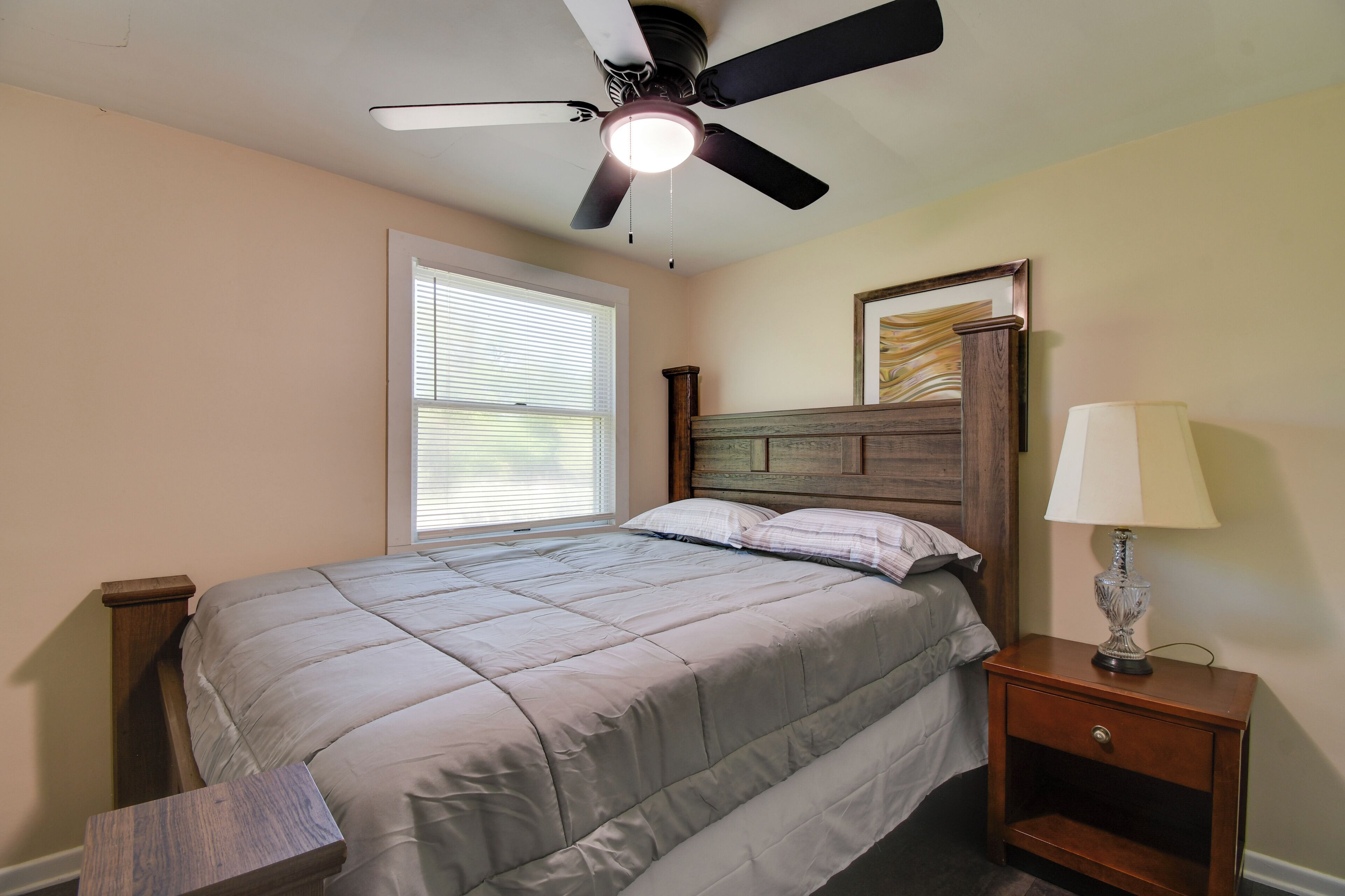 A bedroom with a bed, a nightstand, a lamp, a ceiling fan, and a window with blinds.