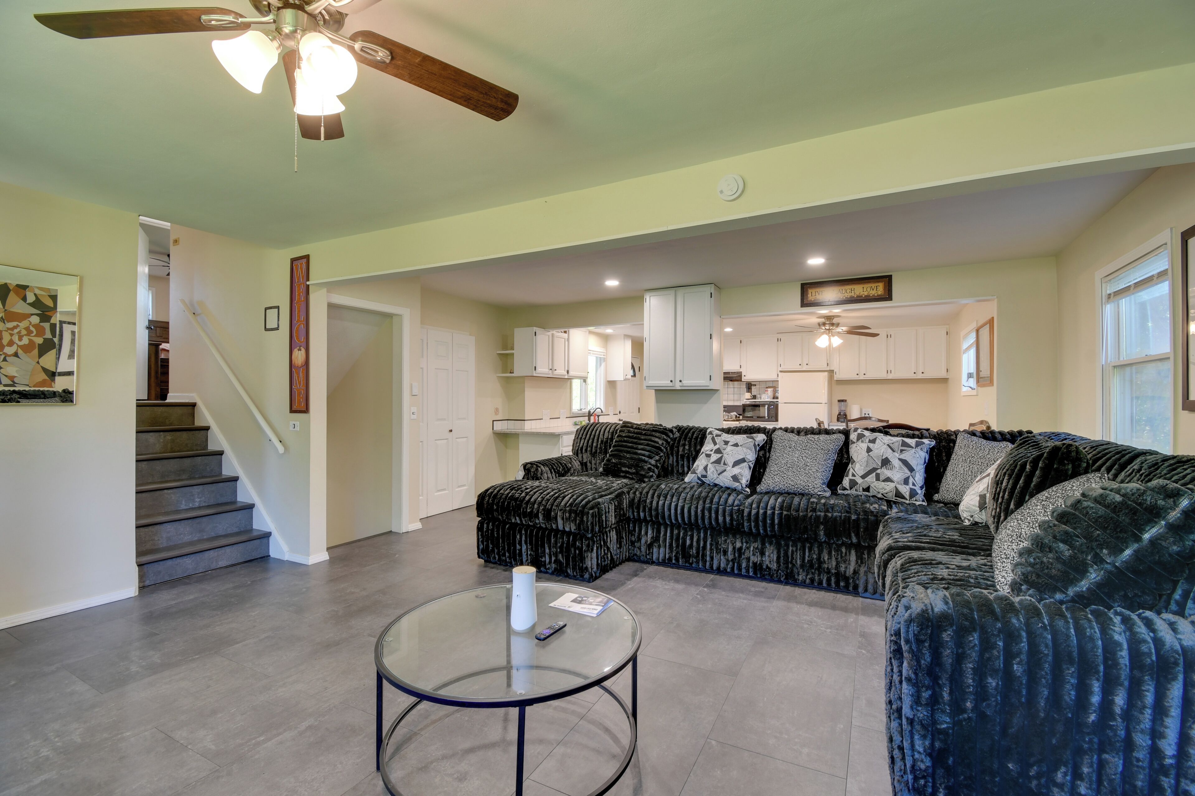 A living room with a sectional sofa, a glass coffee table, a ceiling fan, and a staircase leading to a kitchen.