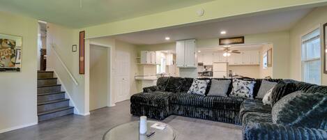 A living room with a sectional sofa, a glass coffee table, a ceiling fan, and a staircase leading to a kitchen.