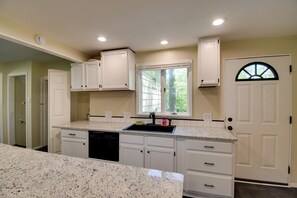 A kitchen with white cabinets, a granite countertop, a dishwasher, and a window with a view of trees.