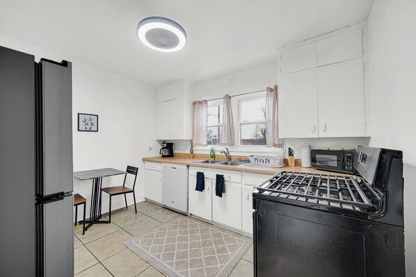 A compact kitchen with white cabinets, a black stove, a microwave, a sink, and a small dining table with chairs. - Superhost: 2BR Dtwn Apt Extended Stay Welcome (Michigan City)
