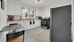 Et lille køkken med hvide skabe, en mikrobølgeovn, ovn, køleskab, kaffemaskine og et vindue med gardiner.