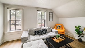 A room with a sectional sofa, a small table, an orange armchair, and a potted plant. - Extended-stay Apartment in Downtown MC Near Lake (Michigan City)