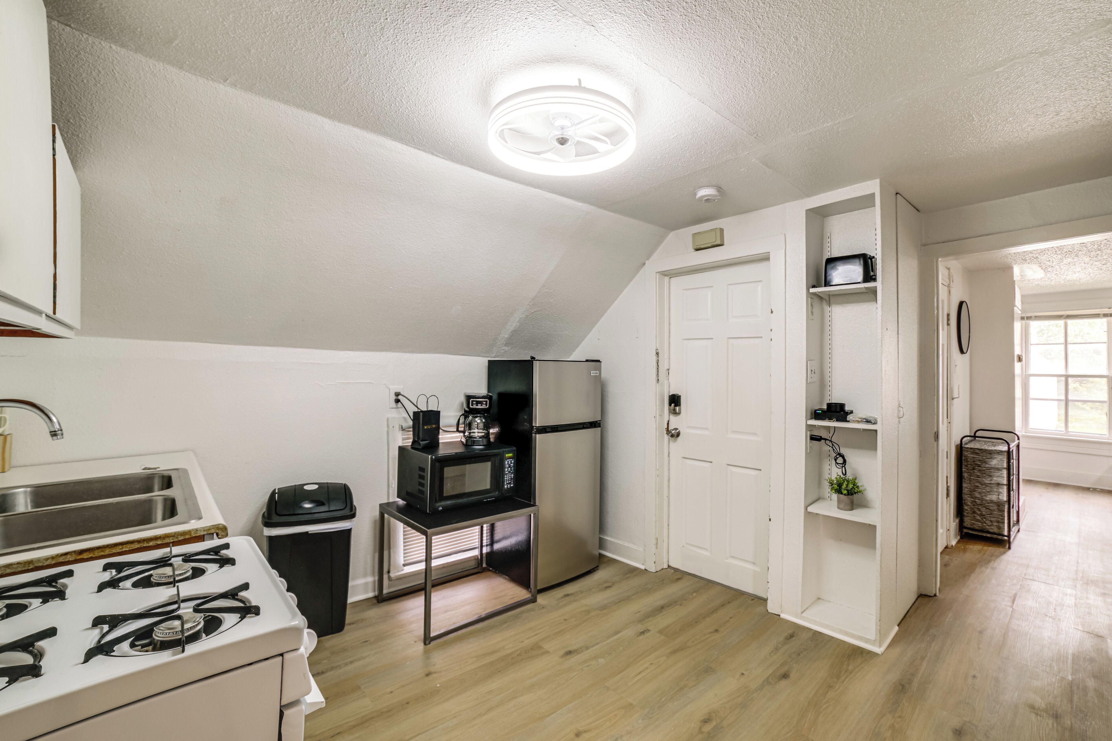 Una cocina compacta con estufa a gas, microondas y refrigerador, un pequeño bote de basura y una puerta blanca que da a otra habitación.