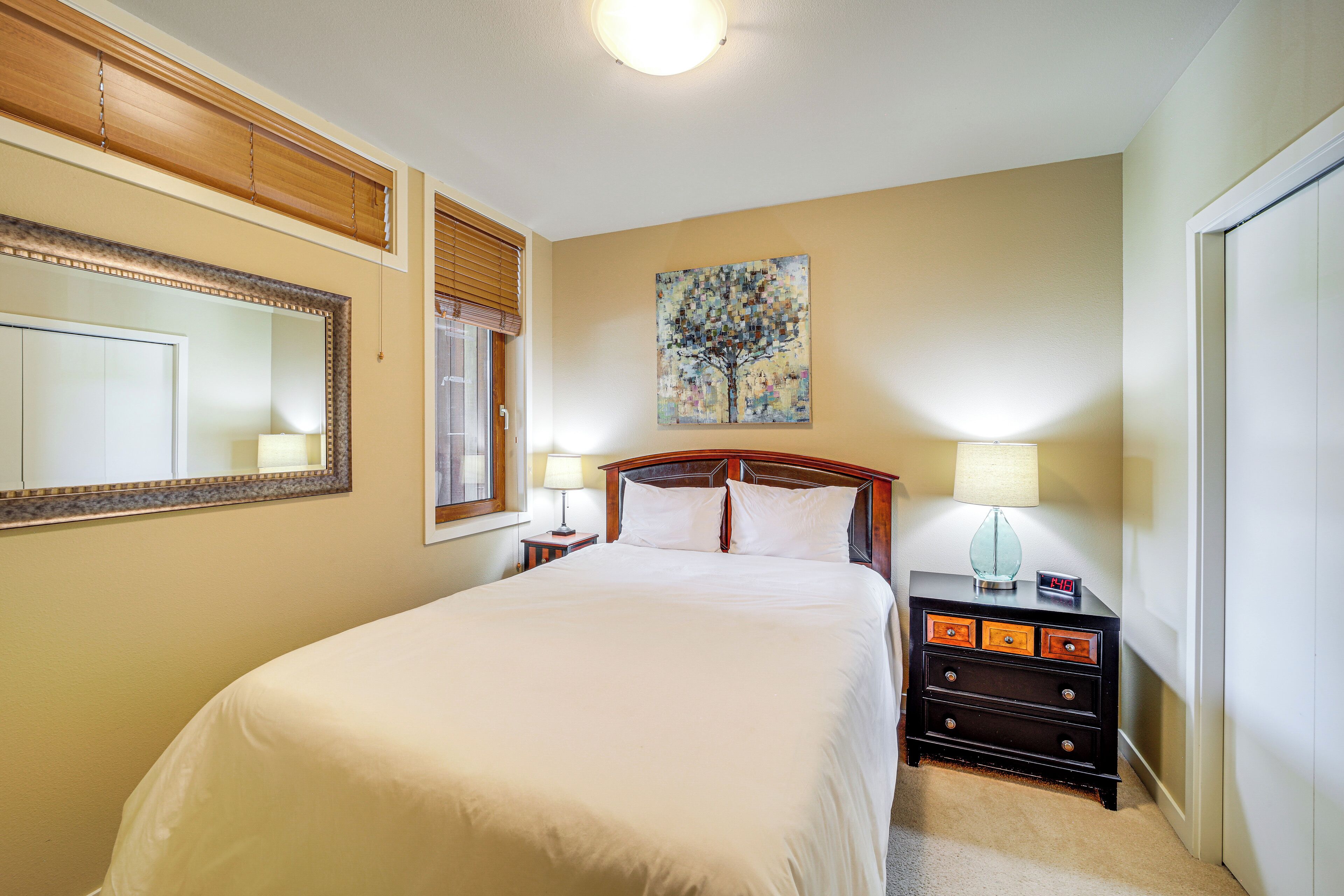 A neatly arranged bedroom with a large bed, a bedside table with a lamp, a nightstand with drawers, a wall-mounted mirror, and a painting of a tree.