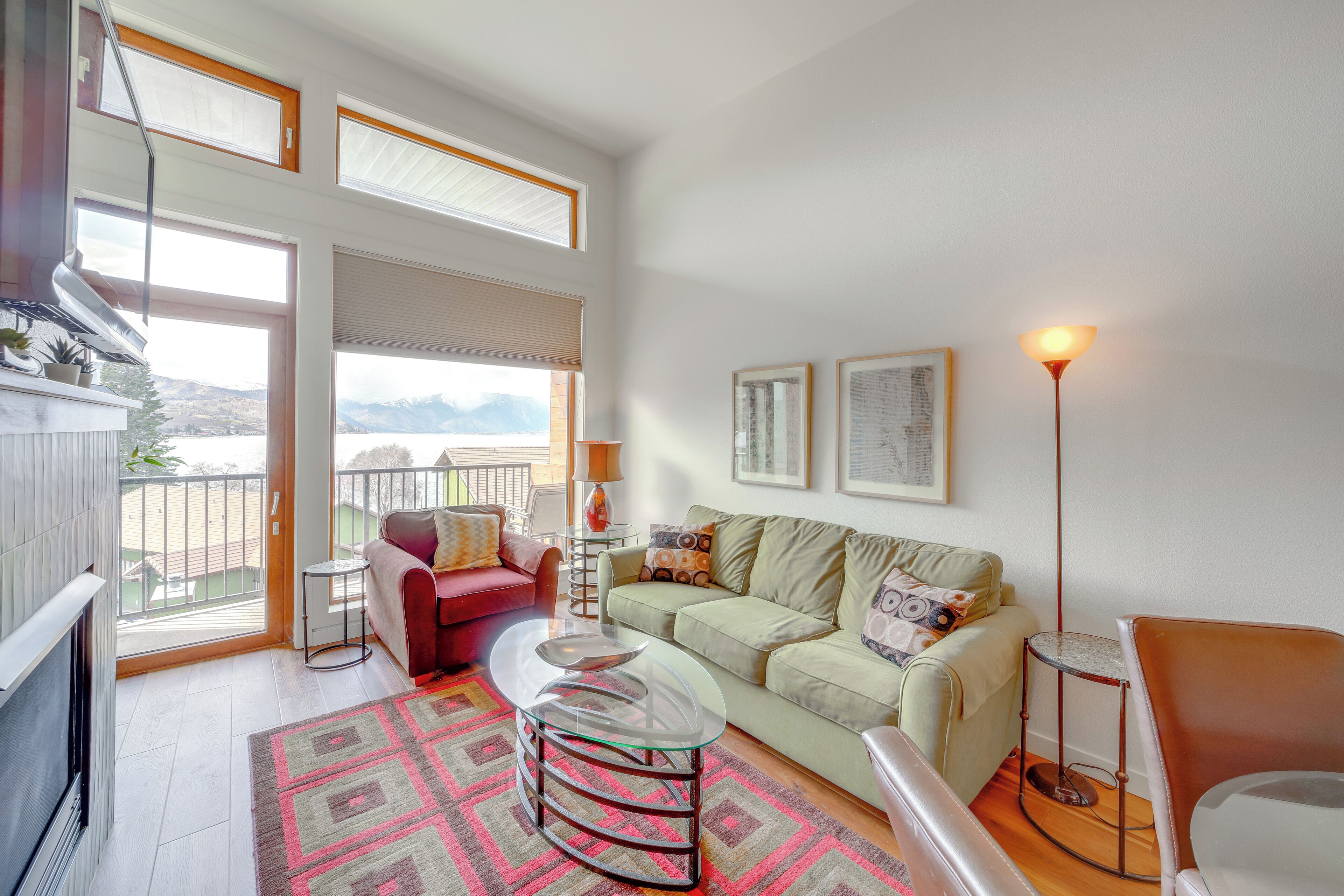 A spacious living area with a sofa, a glass coffee table, and a floor lamp. There is a balcony with a view of the mountains.