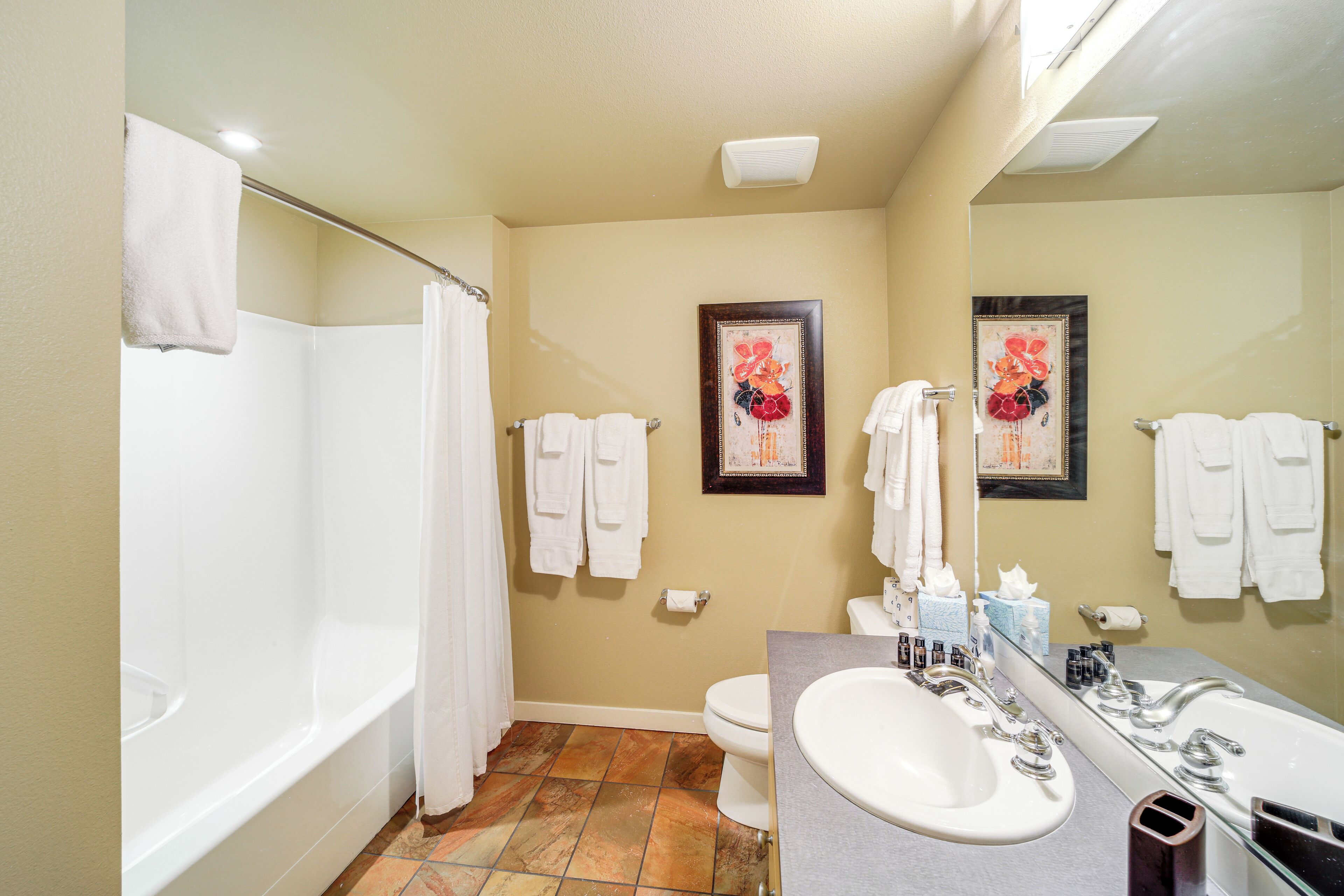 Ein Badezimmer mit Badewanne, Toilette und Waschbecken. Handtücher hängen an der Wand und ein gerahmtes Bild ist über der Toilette angebracht.