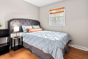 A bedroom with a tufted headboard, a bedside table with a lamp, a window with blinds, and a radiator.