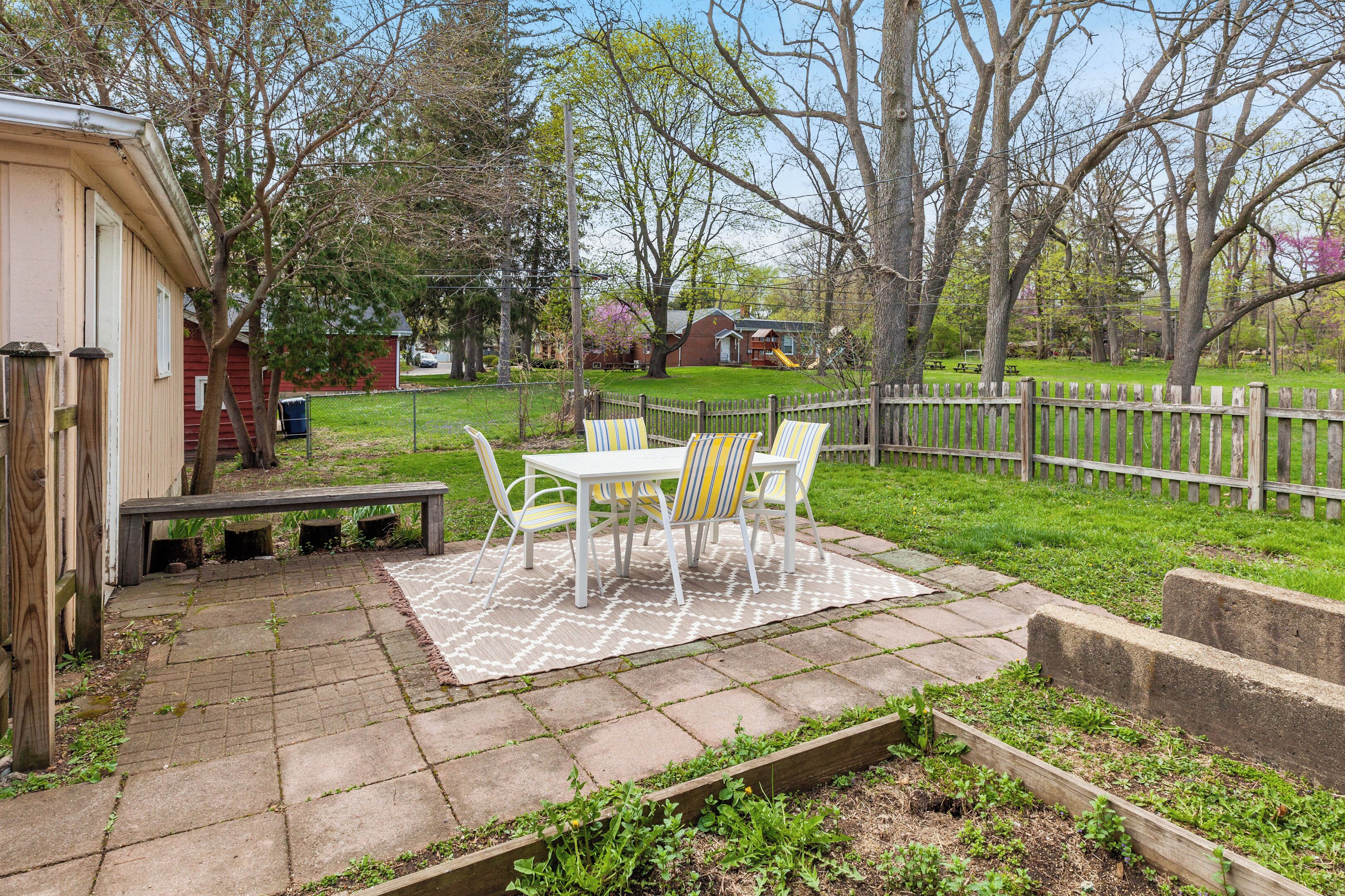 Un patio con una mesa y sillas, un banco de madera y un cantero con plantas.