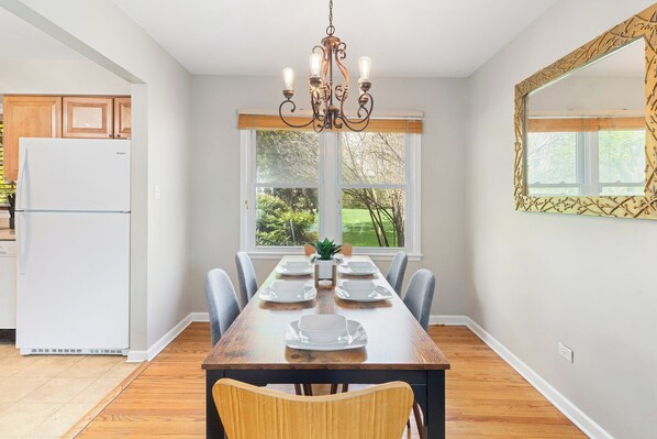 A dining room with a wooden table, chairs, a chandelier, and a view of the outdoors through large windows.