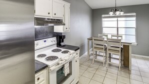 Una cocina con armarios blancos, un refrigerador de acero inoxidable, una estufa blanca y una mesa de comedor con sillas.