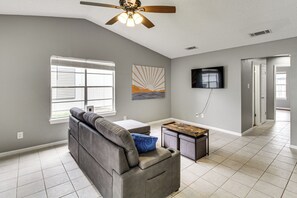 Ein Wohnzimmer mit grauem Sofa, einem Couchtisch, Deckenventilator, Wand-Fernseher und einem Fenster mit Jalousien.