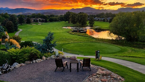 Campo de golf con un estanque, sillas y una mesa en primer plano, y casas al fondo.