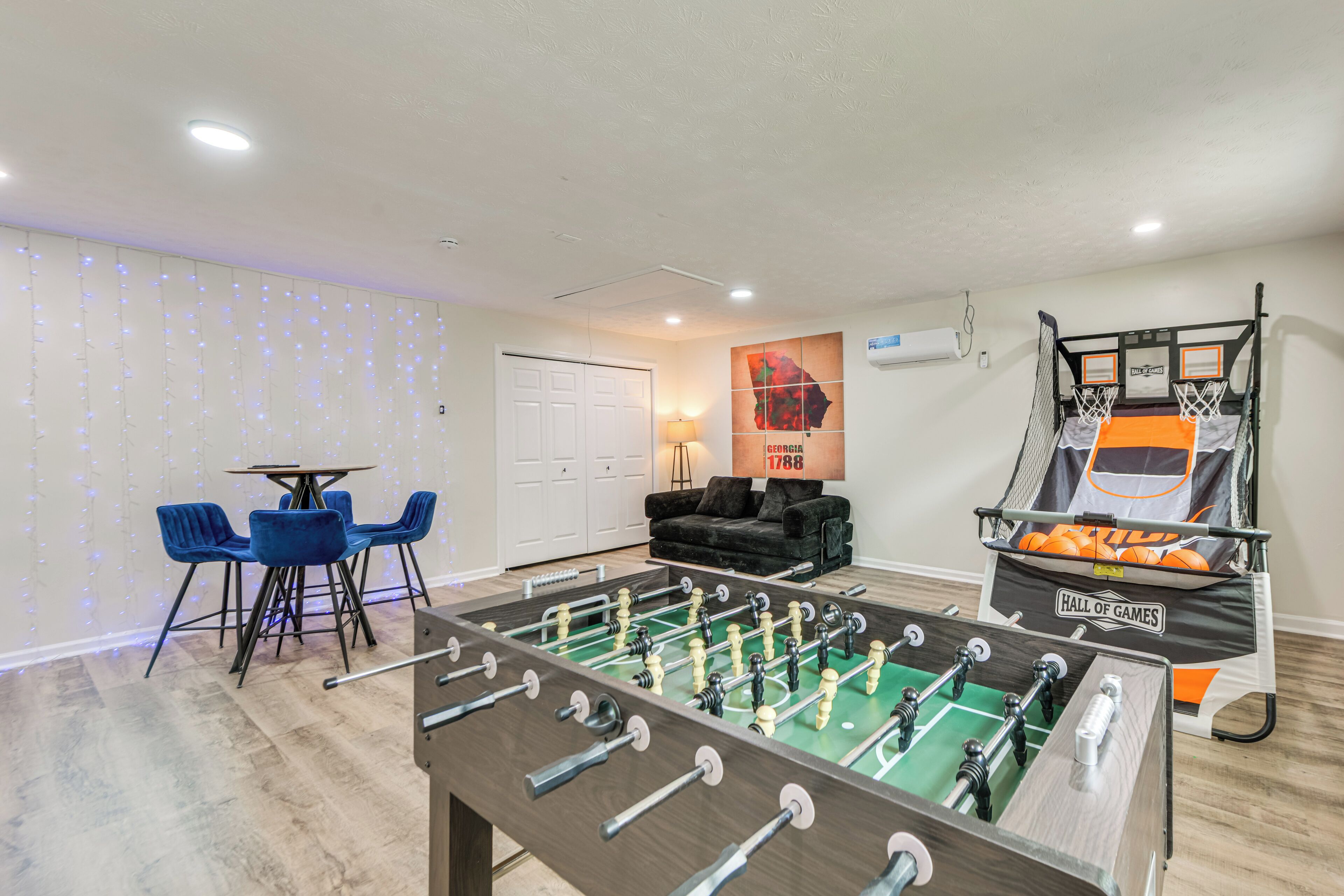 A room with a foosball table, a basketball hoop, and a dining area with blue chairs.