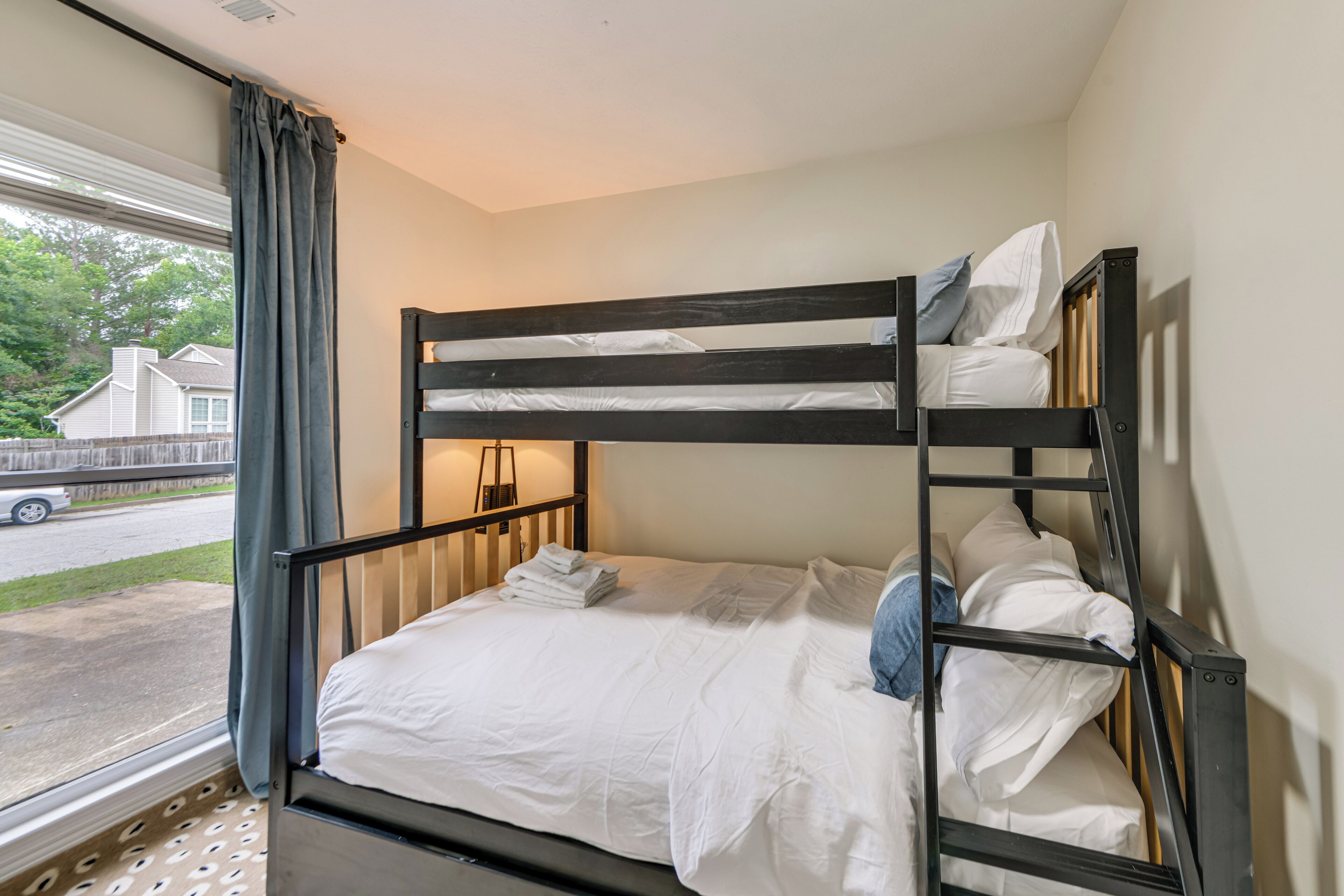 A bunk bed with white bedding, a window with curtains, and a view of a street and houses outside.