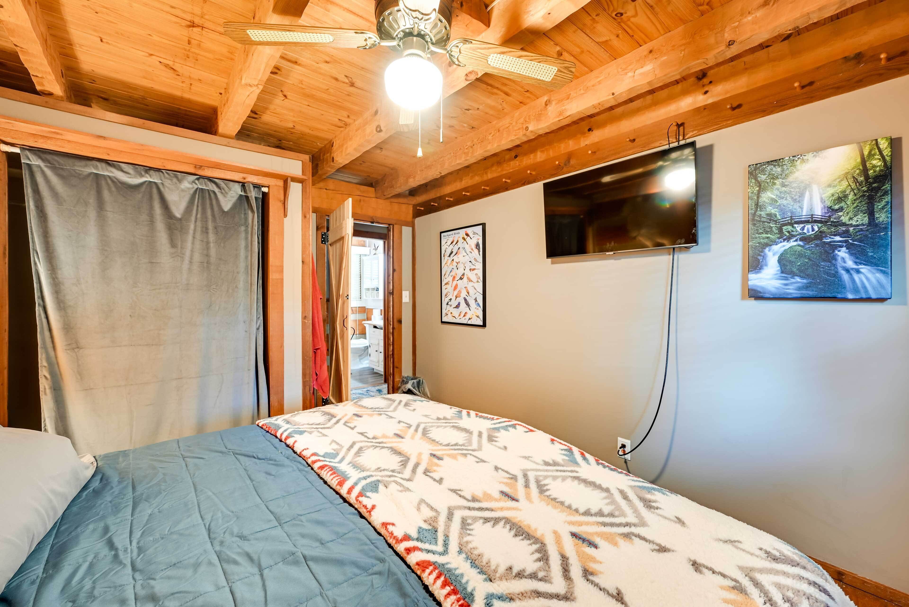 Une chambre avec un plafond en bois, un lit avec une literie à motifs, un ventilateur de plafond et un téléviseur fixé au mur.