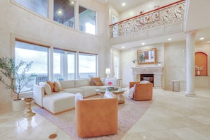 A spacious living area with a high ceiling, large windows, a fireplace, and a balcony.