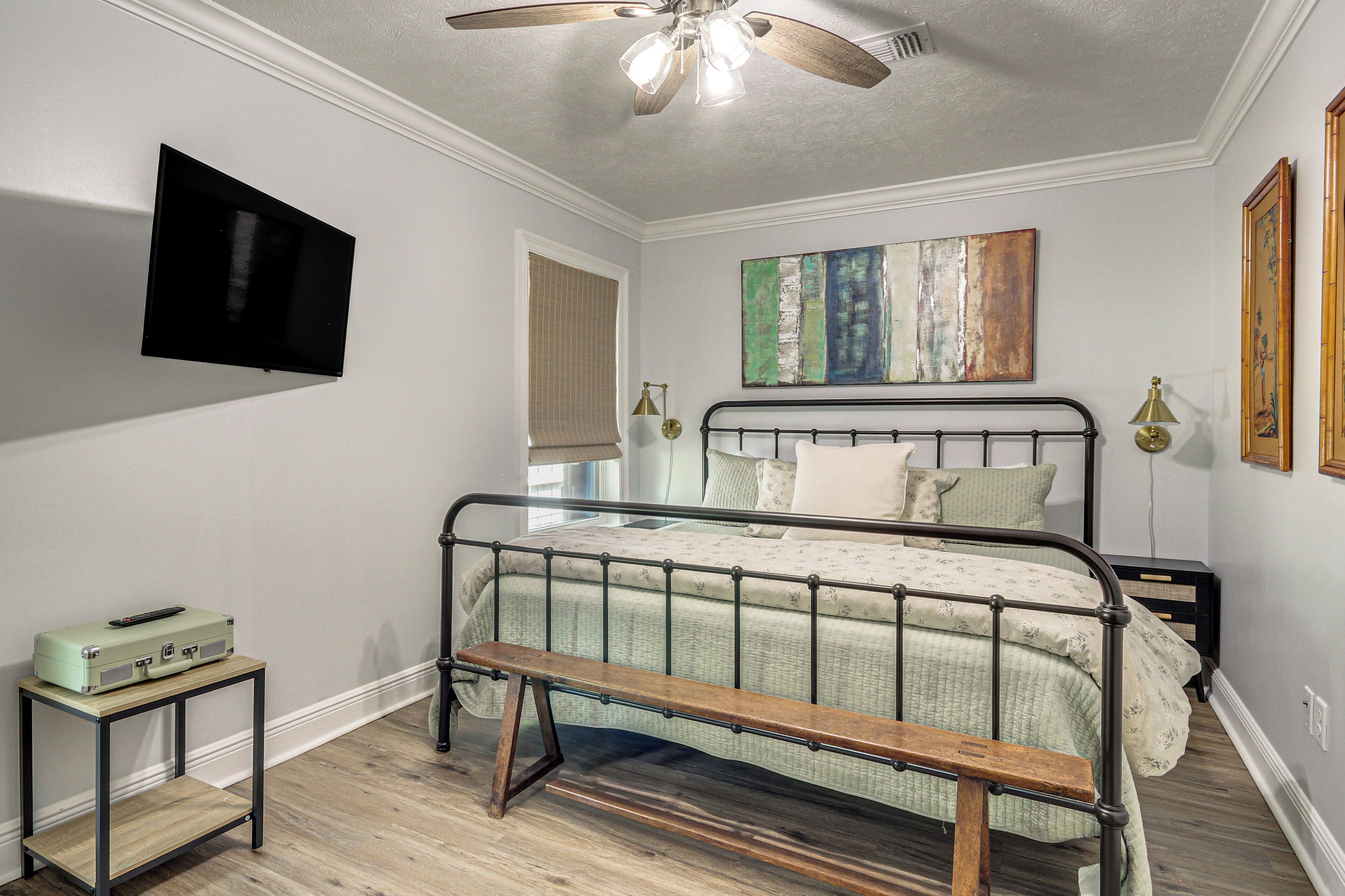 Un dormitorio con cama, ventilador de techo, televisor montado y un cuadro en la pared.