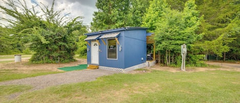 Une petite cabane bleue, dotée d’une porte et de fenêtres, entourée d’arbres et bordée d’un chemin de gravier.