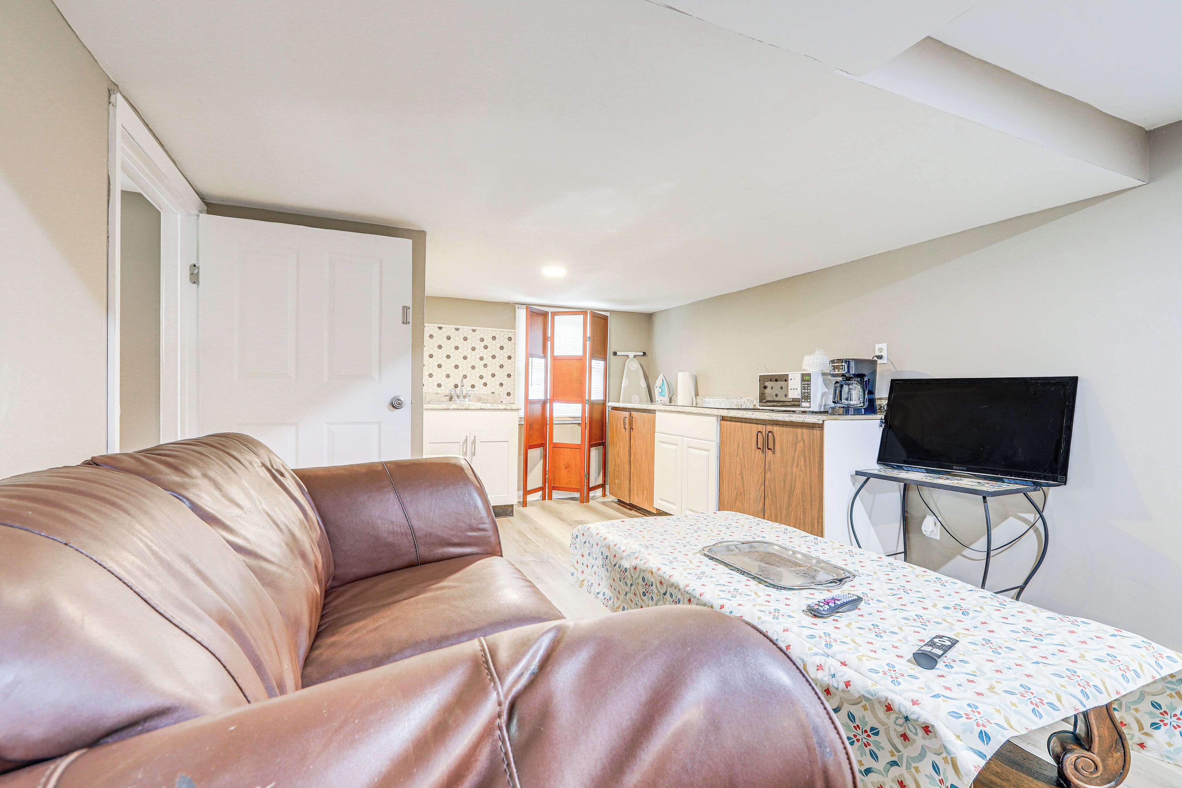 A living room with a brown leather sofa, a small table with a floral tablecloth, a television on a stand, and a kitchen area in the background.