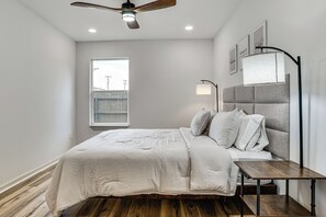 A bedroom with a bed, bedside table, lamp, ceiling fan, window, and artwork on the wall.