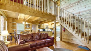 A wooden cabin interior with a leather sofa, a dining area, a kitchen, and a wooden staircase. - Smoky Mtn Oasis w/ Hot Tub! 5 Mi to Skyland Ranch (Sevierville)