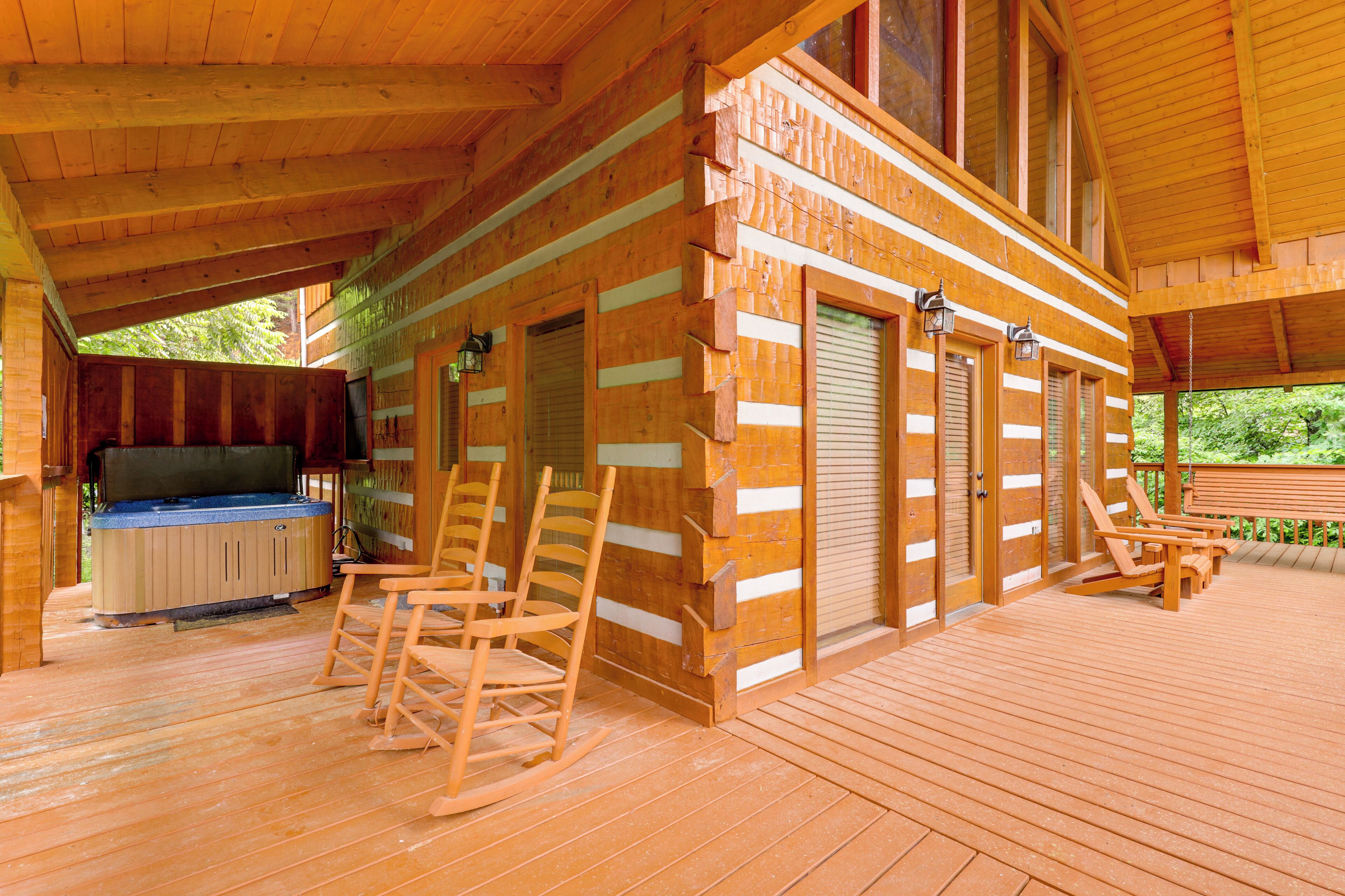 A wooden cabin with a deck, rocking chairs, a hot tub, and a covered patio area.