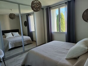 A bedroom with a large mirror, two beds, a hanging light fixture, and a window with curtains. - House in the heart of a vineyard in Provence at the foot of Mont Ventoux (Carpentras)