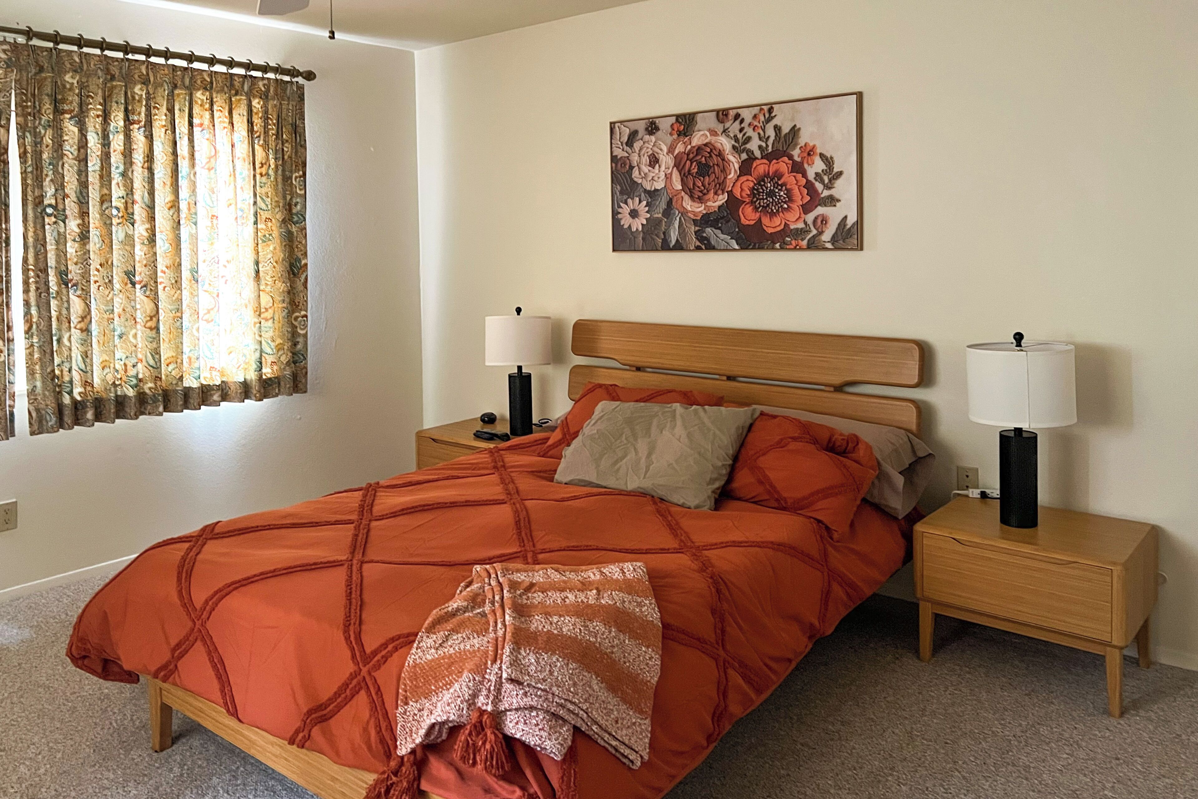 A bedroom with a bed, two bedside tables with lamps, a window with curtains, and a floral painting on the wall.