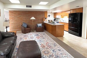 Ein modernes Wohnzimmer mit einem braunen Ledersofa, zwei Sesseln, einem gemusterten Teppich und einer Küchenzeile mit Holzausbau und einem schwarzen Kühlschrank.