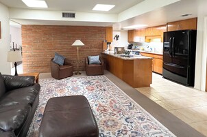 Un salón moderno con un sofá de cuero marrón, dos sillones, un tapete estampado y una zona de cocina con armarios de madera y un refrigerador negro.