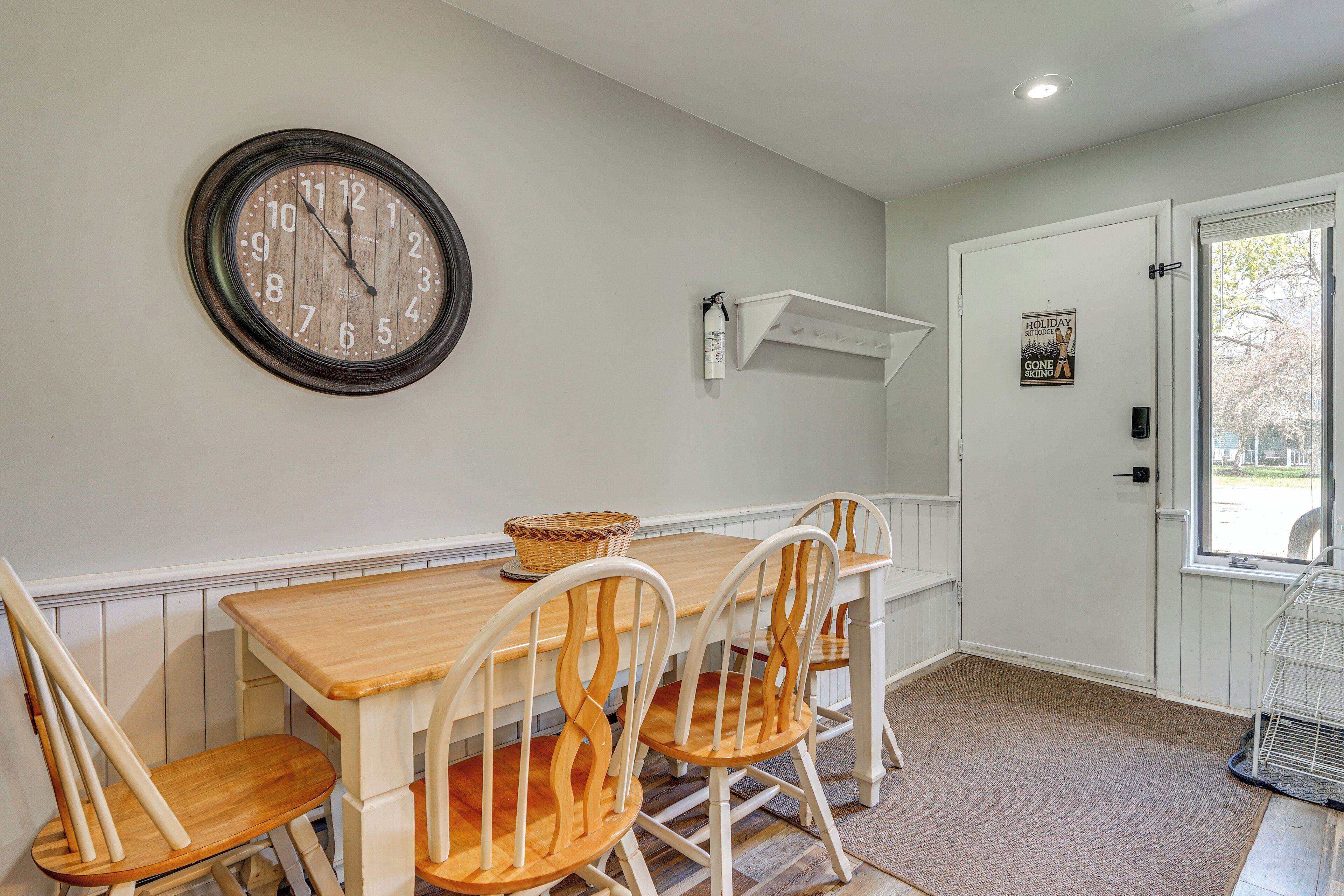 A dining area with a wooden table, four chairs, a wall clock, a white shelf, and a door with a window view.