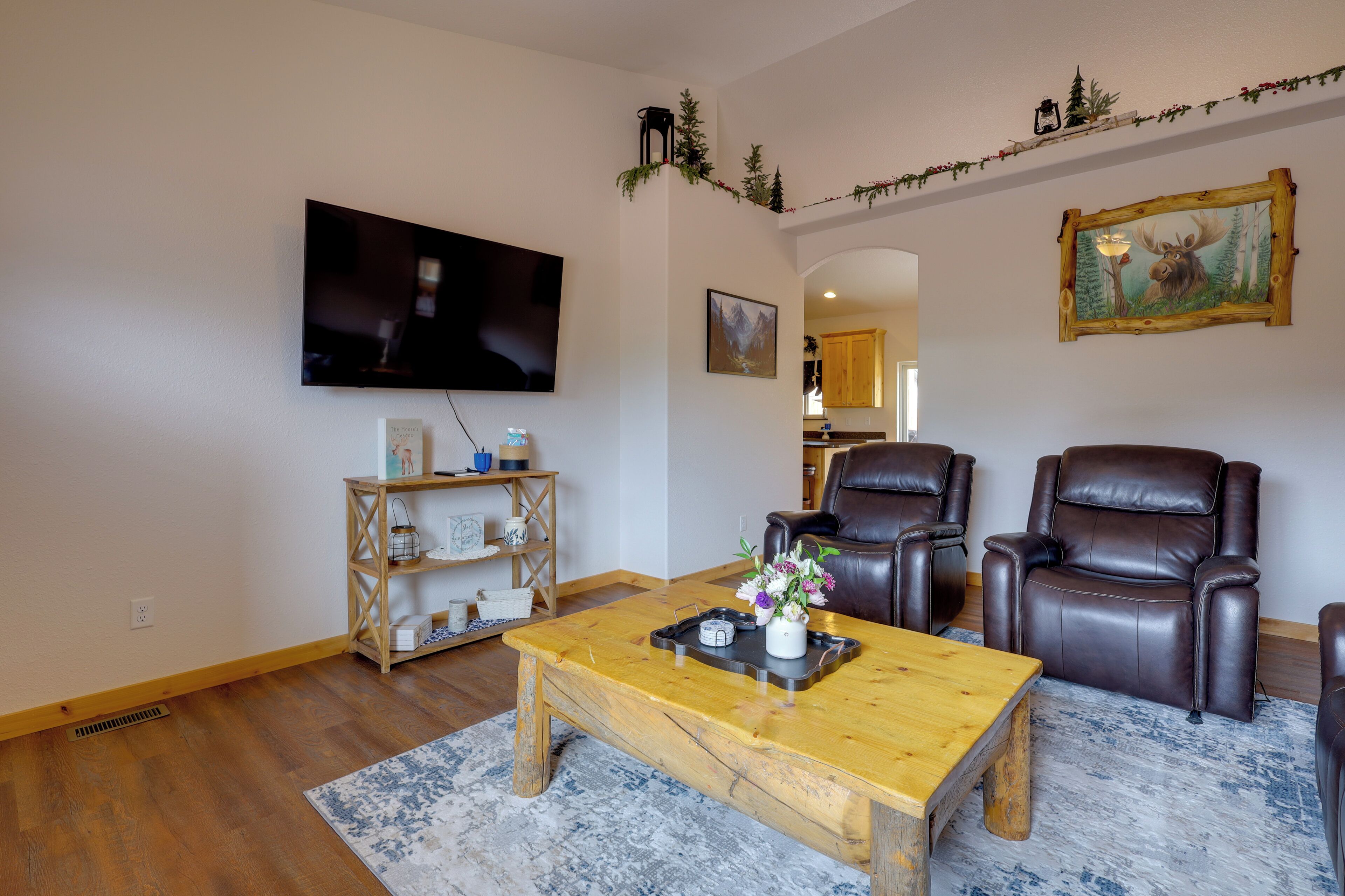 Ein Wohnzimmer mit Flachbildfernseher, Couchtisch aus Holz, zwei Ledersessel, Bücherregal und einem Bild eines Elches.