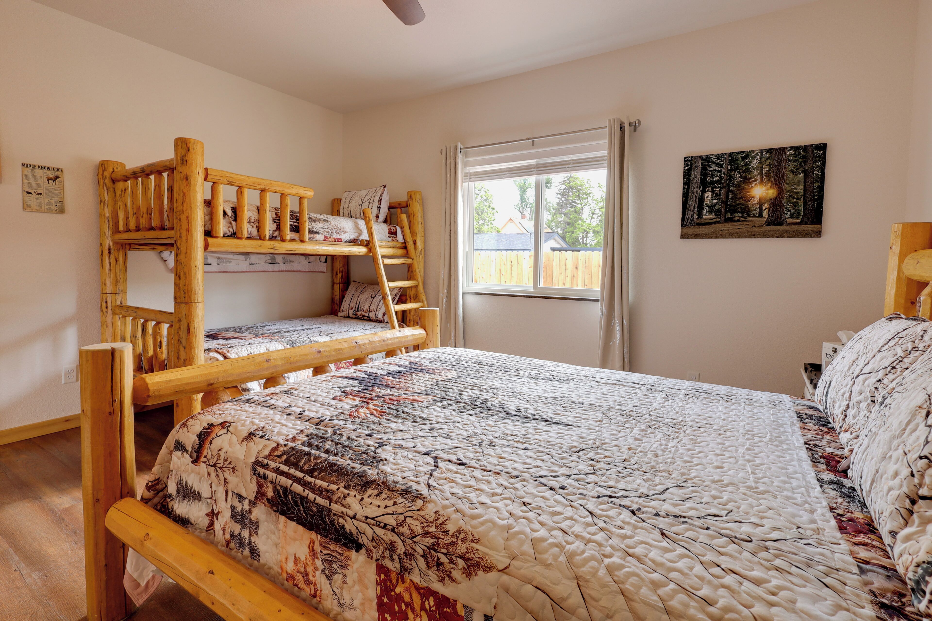 A bunk bed with a forest-themed quilt, a window with a view of a wooden fence, and a forest painting on the wall.