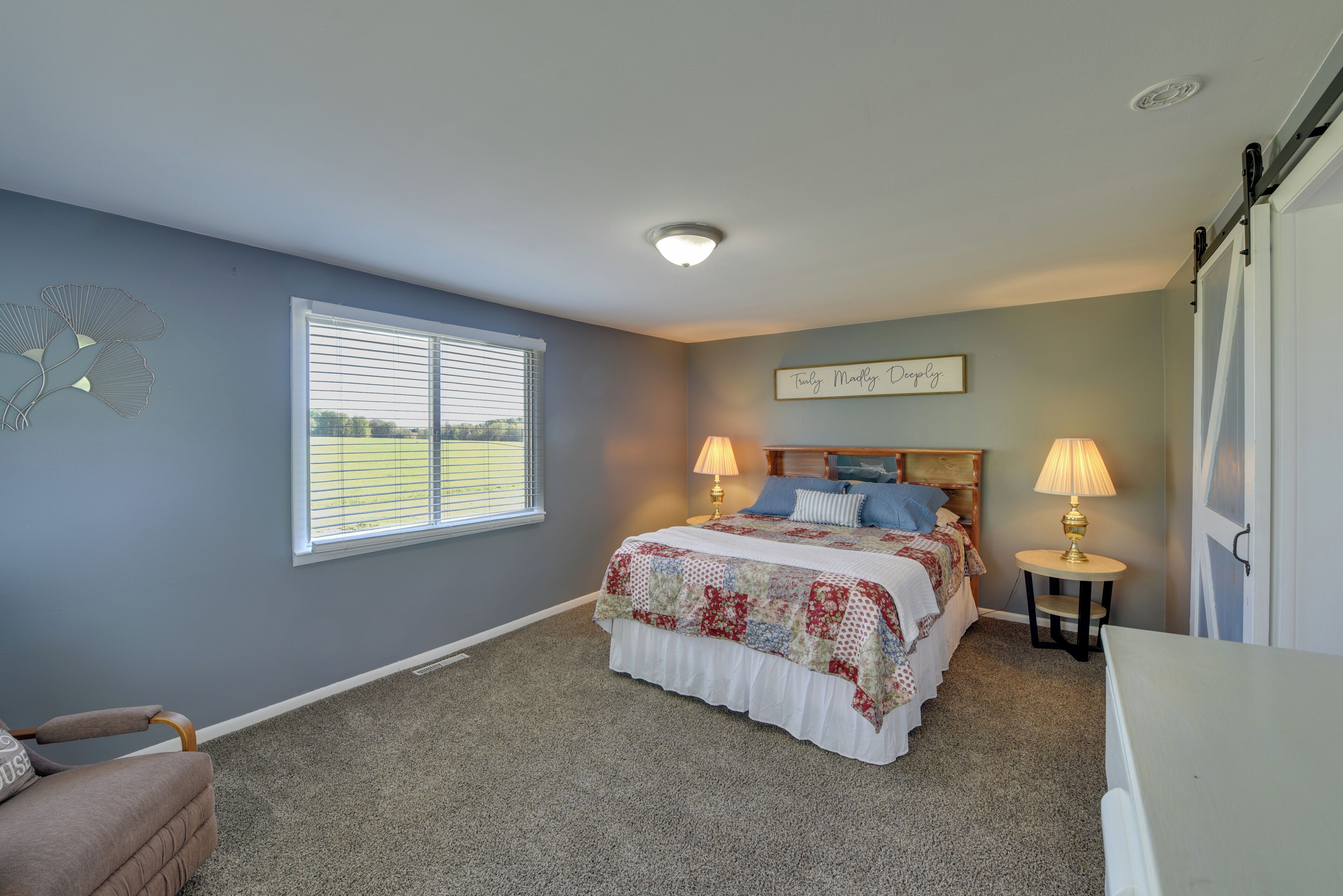 A bedroom with a bed, two bedside lamps, a window with blinds, a chair, and a wall decoration.