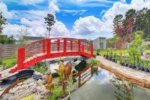Uma ponte vermelha sobre um lago com plantas em vasos e uma cerca de madeira num jardim.
