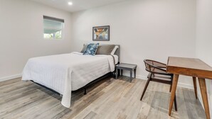 Quarto com cama, mesa pequena, cadeira, ventoinha no teto, janela e um quadro na parede.